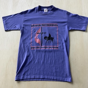 May include: A purple short-sleeved t-shirt featuring a graphic print with a black cross, a silhouette of a person on horseback, and a pink flame. The text reads UNITED METHODISTS and INTO THE FUTURE BY FAITH in black capital letters.