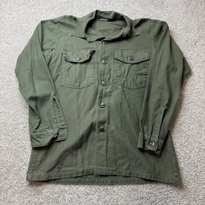 Puede incluir: Camisa verde oliva con botones, dos bolsillos en el pecho y puños abotonados. La camisa de manga larga tiene cuello y cierre de botones. La camisa está hecha de una tela duradera.