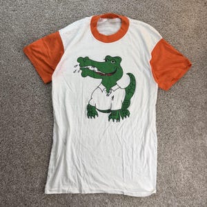 May include: A white t-shirt with orange short sleeves and an orange crew neckline. The center of the shirt features a graphic illustration of a green alligator wearing a white polo shirt, with a small stick figure icon printed on the alligator's shirt.