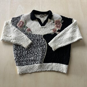 Puede incluir: Un suéter de punto de manga larga con un diseño de bloques de color en negro y blanco con un acabado texturizado. El suéter tiene un cuello tipo polo negro, un escote en V y motivos florales bordados en tonos rosa y verde en el pecho.