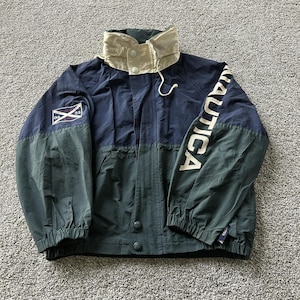 May include: A Nautica jacket featuring a color-blocked design in navy blue and dark green. The jacket has a high beige collar with snap buttons, a front zipper closure, and elastic cuffs. The word NAUTICA is printed in large white letters on the sleeve.