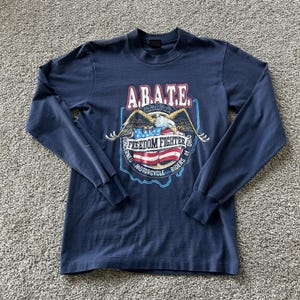 May include: A navy blue long-sleeve t-shirt featuring a graphic print of a bald eagle perched on an American flag. The text reads A.B.A.T.E. C.M.R.O. FREEDOM FIGHTER CONCERNED MOTORCYCLE RIDERS OF OHIO, set against a light blue outline of Ohio.