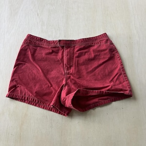May include: A pair of solid dark red shorts featuring a flat waistband, a centre front fly closure, and visible topstitching along the seams. The shorts have a simple, structured design with a straight hemline and a matte fabric finish.