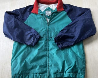 Peut inclure: Un coupe-vent zippé avec un corps sarcelle, des manches raglan bleu marine et un col doublé en polaire rouge. La veste présente une doublure intérieure blanche et une fermeture éclair centrale. Un écusson circulaire avec la lettre C est visible.