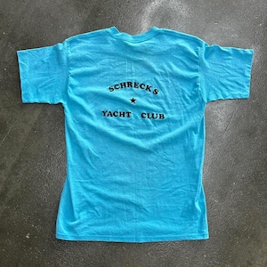 Puede incluir: Una camiseta de manga corta de color turquesa brillante con el texto negro SCHRECK'S YACHT CLUB impreso en la parte posterior, con una pequeña estrella negra centrada entre las dos líneas de texto. La prenda tiene cuello redondo y dobladillo recto.