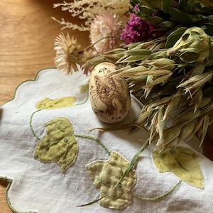 May include: A wooden egg featuring a cat illustration, placed on a white cloth with green leaf embroidery. Dried flowers and foliage are in the background. The egg is a light brown colour.