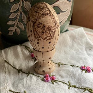 May include: A wooden egg featuring a ferret illustration, displayed in a matching egg cup. The egg is adorned with vertical lines and starbursts. The egg cup sits on a white cloth with pink floral embroidery, with a green ceramic pot in the background.