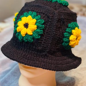 Op de afbeelding: Een zwarte gehaakte bucket hat met meerdere driedimensionale bloemmotieven. Elke bloem is opgebouwd met een felgeel hart omringd door donkergroene bloemblaadjes. De hoed is gemaakt van donker garen met een brede, naar beneden lopende rand.