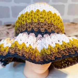 May include: A crochet bucket hat featuring a wavy, chevron-style pattern in horizontal stripes of white, olive green, brown, and dark navy blue. The hat has a rounded crown and a wide, flexible brim with a textured, hand-stitched yarn construction.