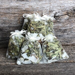 May include: Five small white mesh bags filled with dried herbs and white flower petals. The bags are arranged on a rustic wooden surface.