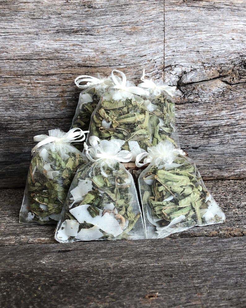 May include: Five small white mesh bags filled with dried herbs and white flower petals. The bags are arranged on a rustic wooden surface.