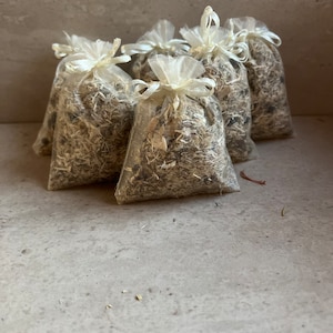 May include: A group of six small, white, mesh bags filled with dried herbs and tied with white ribbon. The bags are arranged in a semi-circle on a light gray surface.