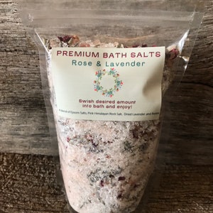 May include: A clear plastic bag filled with white bath salts, pink Himalayan rock salt, dried lavender, and rose petals. The bag has a white label that reads "PREMIUM BATH SALTS Rose & Lavender Swish desired amount into bath and enjoy! A blend of Epsom Salts, Pink Himalayan Rock Salt, Dried Lavender and Roses."