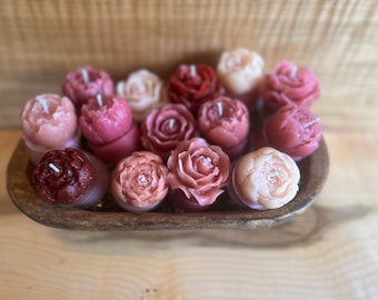 May include: A collection of small, pink, red, and white floral-shaped candles in a wooden bowl. The candles are arranged in a gradient of color, with the darkest red candles at the bottom and the lightest white candles at the top.