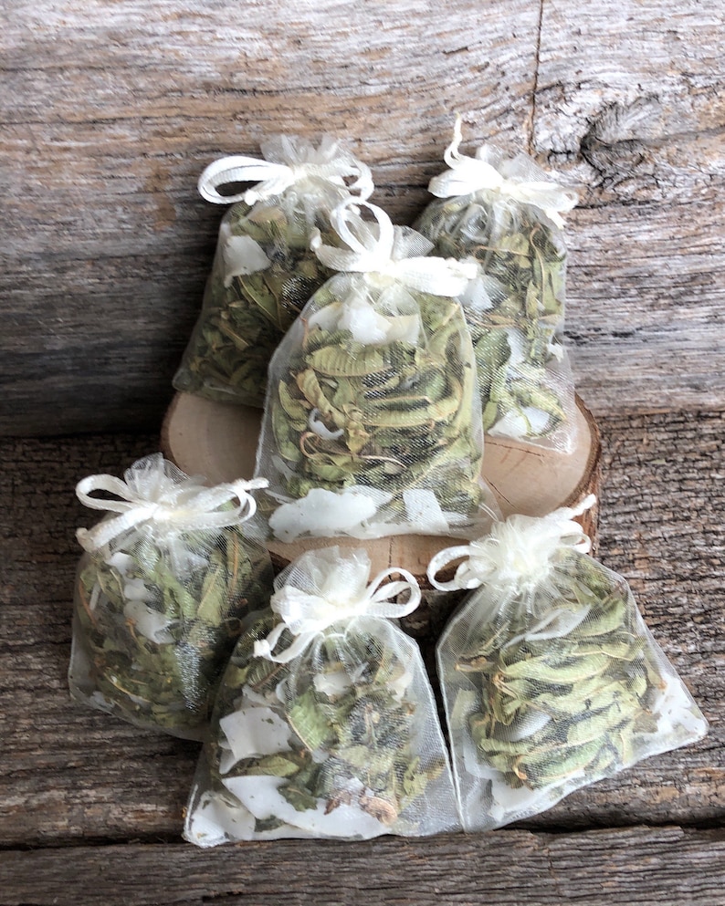 May include: Six small white organza bags filled with dried herbs. The bags are tied with white ribbon and are arranged on a wooden surface.
