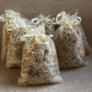 May include: Four small white mesh bags filled with dried herbs and flowers. The bags are tied with white ribbon and are arranged in a row on a light gray surface.