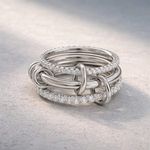 May include: A silver ring with a unique design. The ring features multiple bands, some adorned with small, sparkling stones. The bands are intertwined, creating an intricate and eye-catching pattern. The ring is set against a neutral background.