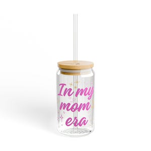 Könnte beinhalten: Ein klares, glasförmiges Trinkglas mit Bambusdeckel und klarem Strohhalm. Auf dem Becher steht in rosa Schrift "In my mom era" mit Stern- und Herzakzenten. Das Design ist auf die Vorderseite des Glases gedruckt.