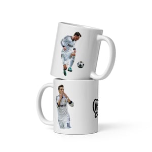 May include: Two white ceramic coffee mugs stacked on top of each other. Each mug features a soccer player in a white uniform. One mug shows a player kicking a soccer ball, and the other shows a player with his hand over his mouth.