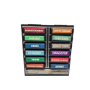 May include: A black, multi-compartment storage unit filled with colorful game cartridges. Each cartridge has a different game title printed on it, including "Barnstorming," "Enduro," "Oink!," "Ice Hockey," "Tennis," "Pressure Cooker," "Chopper Command," "Checkers," "Robot Tank," "Dragster," "Boxing/La Boxe," and "Skiing."