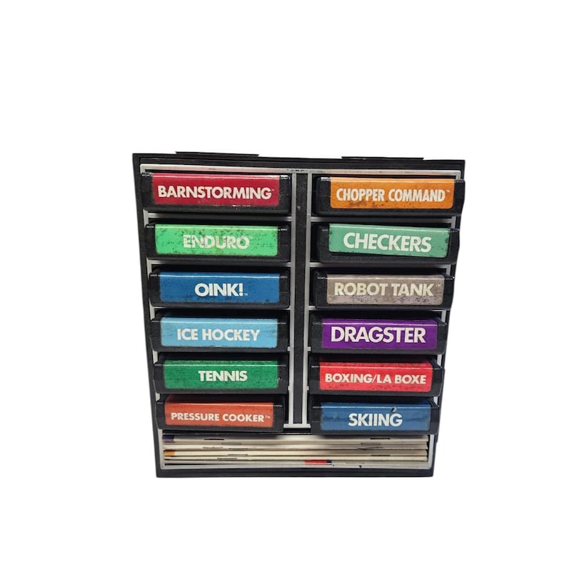 May include: A black, multi-compartment storage unit filled with colorful game cartridges. Each cartridge has a different game title printed on it, including "Barnstorming," "Enduro," "Oink!," "Ice Hockey," "Tennis," "Pressure Cooker," "Chopper Command," "Checkers," "Robot Tank," "Dragster," "Boxing/La Boxe," and "Skiing."