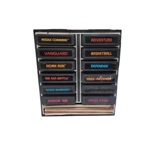 Puede incluir: Un estante negro con cartuchos de videojuegos Atari. Los cartuchos están etiquetados con títulos de juegos como "Missile Command", "Vanguard" y "Space Invaders". También hay una pila de manuales de juegos.