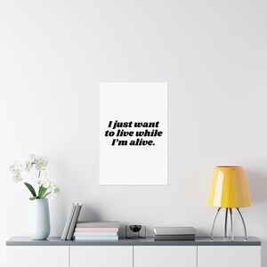 May include: A rectangular white wall poster featuring the black text "I just want to live while I'm alive." centred in a bold, serif font. The poster is displayed on a plain wall above a surface with a white vase of flowers and a yellow lamp.