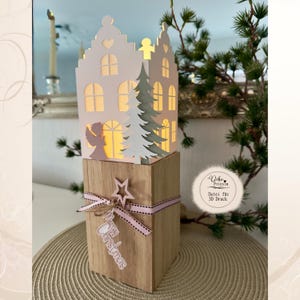 May include: A white paper house lantern with cut-out windows and an angel silhouette, illuminated from within. It sits atop a square wooden block wrapped with a pink-dotted ribbon, a wooden star, and a white Merry Christmas tag.