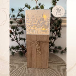 May include: A rectangular wooden block base with a light-up white panel featuring a laser-cut winter village scene with houses, trees, and a streetlamp. A jute twine wraps around the wood with a small wooden tag labeled "leuchtende Weihnachten".