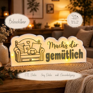 May include: A white rectangular illuminated sign with rounded edges featuring a line art illustration of a sofa with pillows and a sleeping cat. The text reads "Mach's dir gemütlich" in a dark script and sans-serif font with integrated warm lights.