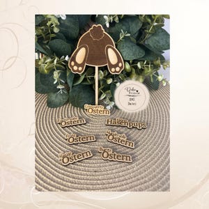 Puede incluir: Decoraciones de Pascua de madera. La imagen presenta una cola de conejo marrón sobre un palo, junto con varios pequeños recortes de madera con la palabra "Ostern" y otras frases alemanas. Las decoraciones están dispuestas sobre una superficie texturizada de color beige.