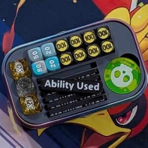 May include: A rectangular, iridescent metal container holding game accessories. It contains dice, counters with numbers, and a green and white circular marker. The text "Ability Used" is visible on a black strip. The background features a colorful illustration.