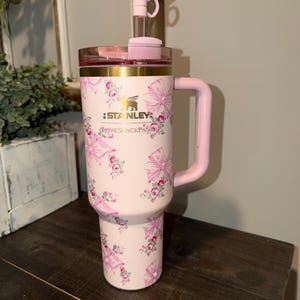 Puede incluir: Un vaso Stanley rosa con un dise&ntilde;o floral y lazos. El vaso tiene un asa rosa, una tapa rosa y un lazo rosa en la pajita. El texto "STANLEY" y "LOVESHACKFANCY" est&aacute;n impresos en el vaso.