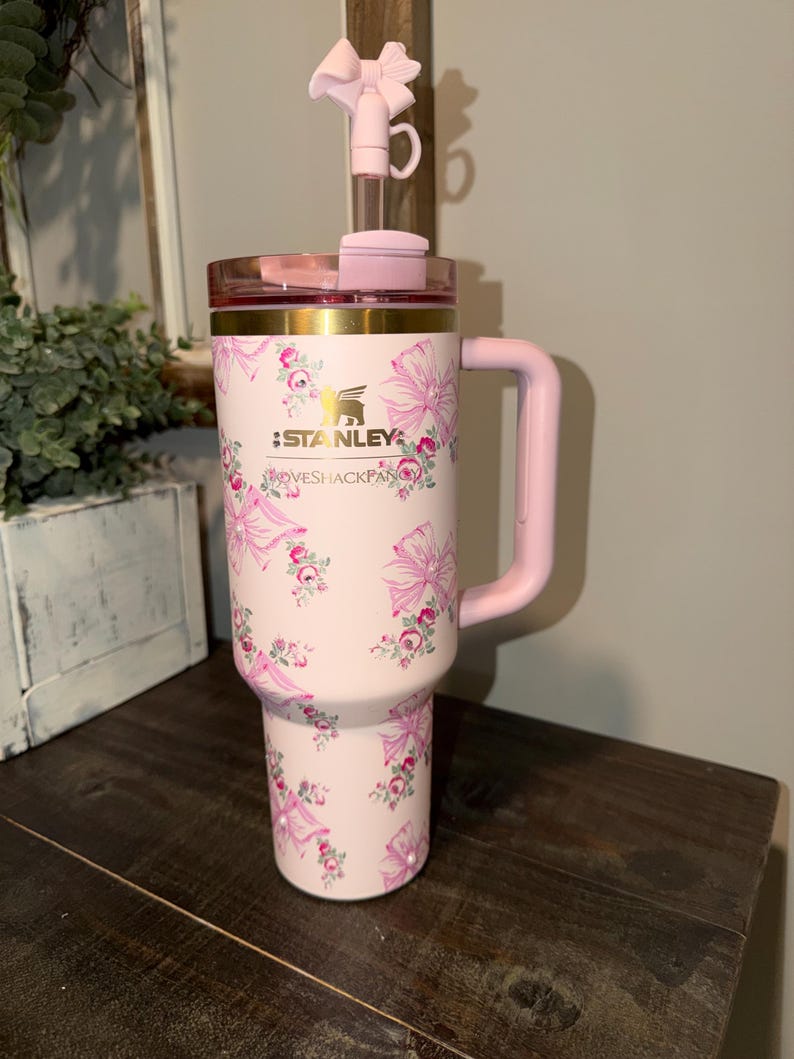 Puede incluir: Un vaso Stanley rosa con un dise&ntilde;o floral y lazos. El vaso tiene un asa rosa, una tapa rosa y un lazo rosa en la pajita. El texto "STANLEY" y "LOVESHACKFANCY" est&aacute;n impresos en el vaso.