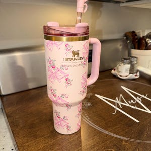Puede incluir: Un vaso Stanley rosa con un dise&ntilde;o floral y lazos. Tiene un asa, tapa y pajita rosas con un lazo decorativo. El logotipo de Stanley es visible. El vaso est&aacute; sobre una superficie de madera.