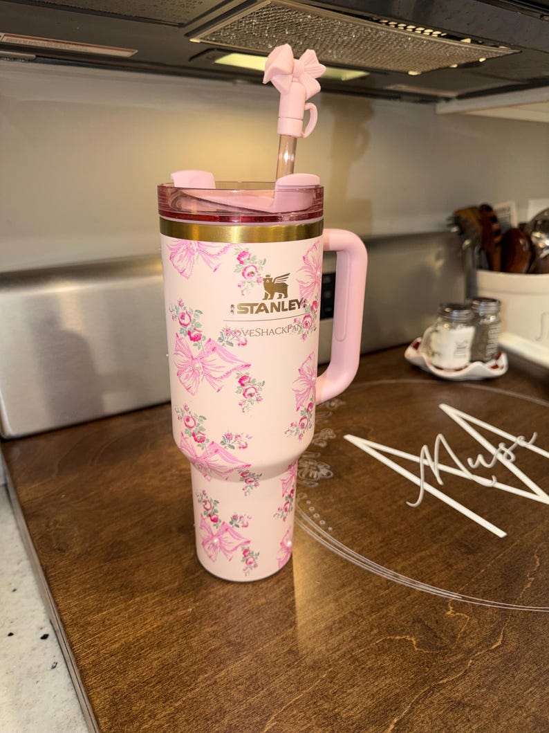 Puede incluir: Un vaso Stanley rosa con un dise&ntilde;o floral y lazos. Tiene un asa, tapa y pajita rosas con un lazo decorativo. El logotipo de Stanley es visible. El vaso est&aacute; sobre una superficie de madera.