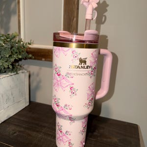 Puede incluir: Un vaso Stanley rosa con un estampado floral y lazos. El vaso tiene un asa, tapa y pajita rosas con un lazo. El texto "LOVESHACKFANCY" est&aacute; impreso en el vaso.