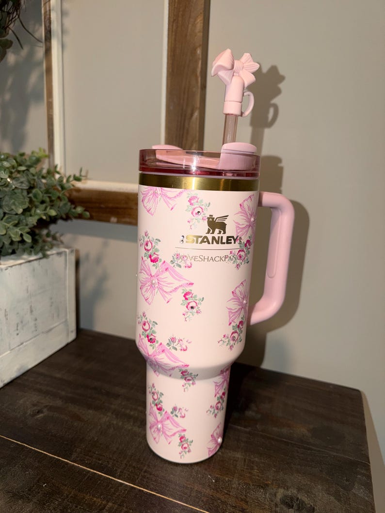 Puede incluir: Un vaso Stanley rosa con un estampado floral y lazos. El vaso tiene un asa, tapa y pajita rosas con un lazo. El texto "LOVESHACKFANCY" est&aacute; impreso en el vaso.
