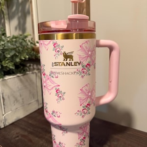 Puede incluir: Un vaso Stanley rosa con un dise&ntilde;o floral y lazos. El vaso tiene un asa, una pajita y una tapa rosa. El logotipo de Stanley y el texto "LOVESHACKFANCY" son visibles.