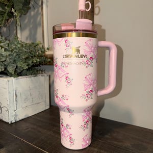 Puede incluir: Un vaso Stanley rosa con un estampado floral y lazos. El vaso tiene un asa y una tapa transparente con un lazo rosa. El texto "STANLEY" es visible.