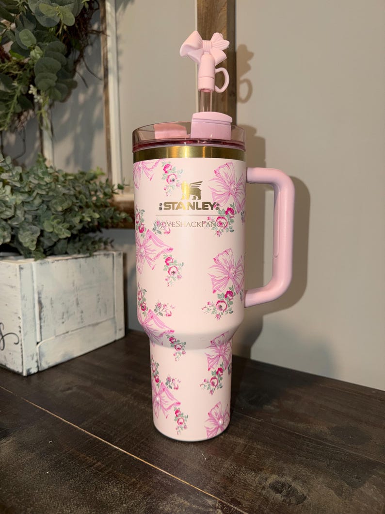 Puede incluir: Un vaso Stanley rosa con un estampado floral y lazos. El vaso tiene un asa y una tapa transparente con un lazo rosa. El texto "STANLEY" es visible.
