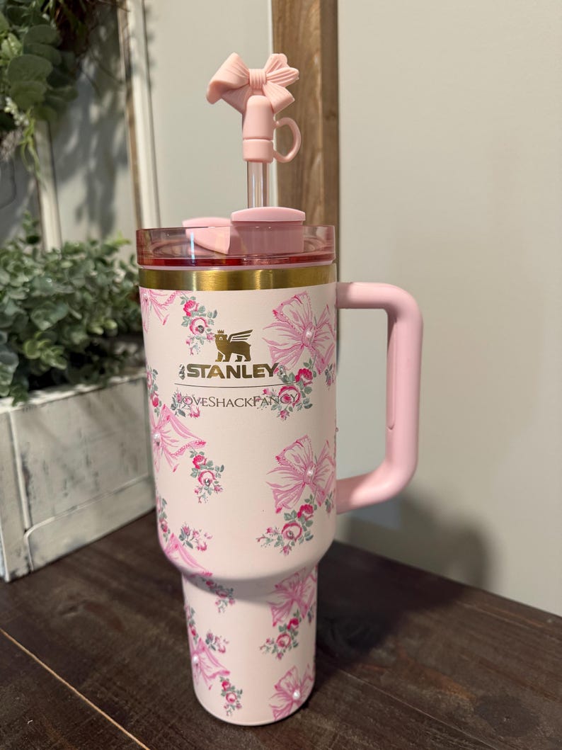 Puede incluir: Un vaso Stanley rosa con un estampado floral y de lazo. Tiene un asa rosa, una tapa transparente con borde dorado y un adorno superior en forma de lazo rosa. El texto "LOVESHACKFANCY" est&aacute; impreso.