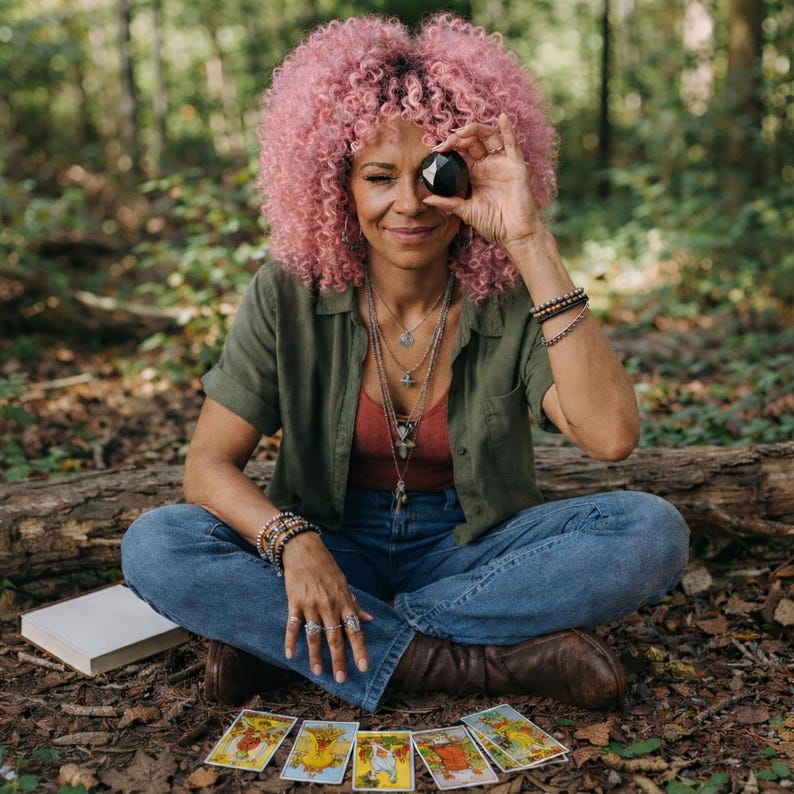 May include: A woman with pink curly hair holds a black crystal over one eye while sitting cross-legged outdoors. She wears a green shirt, red top, and blue jeans. Tarot cards are laid out in front of her, suggesting a spiritual or mystical theme.
