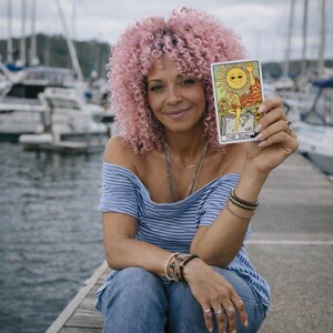 May include: A woman with pink curly hair holds a tarot card depicting the sun. The card features a smiling sun and a figure. She wears a blue and white striped off-the-shoulder top, jeans, and jewelry. The background shows boats and water.