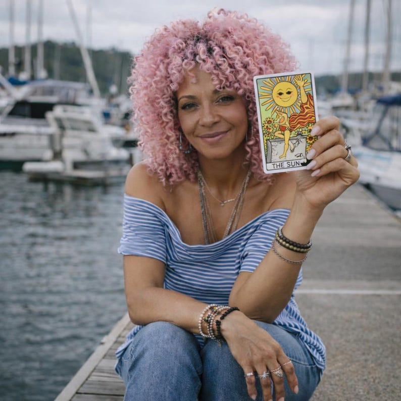 May include: A woman with pink curly hair holds a tarot card depicting the sun. The card features a smiling sun and a figure. She wears a blue and white striped off-the-shoulder top, jeans, and jewelry. The background shows boats and water.