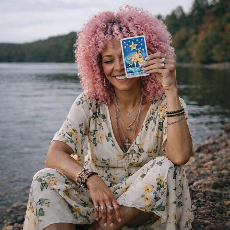 May include: A person with pink curly hair smiles while holding a tarot card in front of their face. The card features a blue background with a star and two figures. They wear a floral dress and multiple necklaces and bracelets.