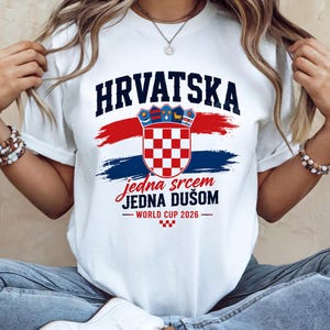 May include: White t-shirt featuring the word "HRVATSKA" in navy blue above a Croatian coat of arms and red and blue brushstrokes. The text "jedna srcem JEDNA DU&Scaron;OM - WORLD CUP 2026" is below the graphic. The shirt is a crew neck.
