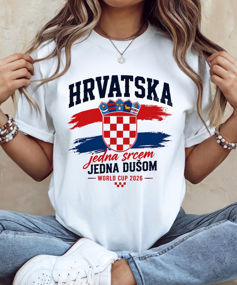 May include: White t-shirt featuring the word "HRVATSKA" in navy blue above a Croatian coat of arms and red and blue brushstrokes. The text "jedna srcem JEDNA DU&Scaron;OM - WORLD CUP 2026" is below the graphic. The shirt is a crew neck.