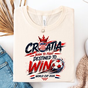 May include: Cream-colored t-shirt with the word "CROATIA" in blue and red, a Croatian coat of arms, and a soccer ball graphic. Text reads "BORN TO FIGHT, DESTINED TO WIN, WORLD CUP 2026."