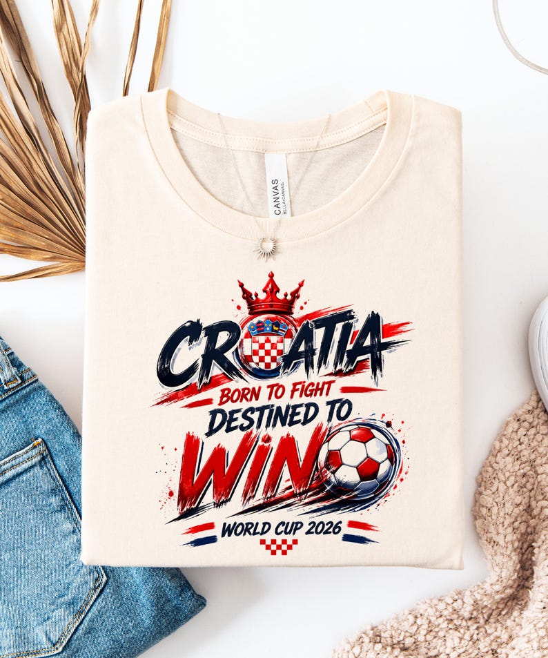 May include: Cream-colored t-shirt with the word "CROATIA" in blue and red, a Croatian coat of arms, and a soccer ball graphic. Text reads "BORN TO FIGHT, DESTINED TO WIN, WORLD CUP 2026."